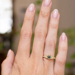 A close-up of a hand wearing a Vintage-inspired, yellow gold engagement ring with a round-cut teal sapphire and six small accent diamonds, set against a softly blurred outdoor background.