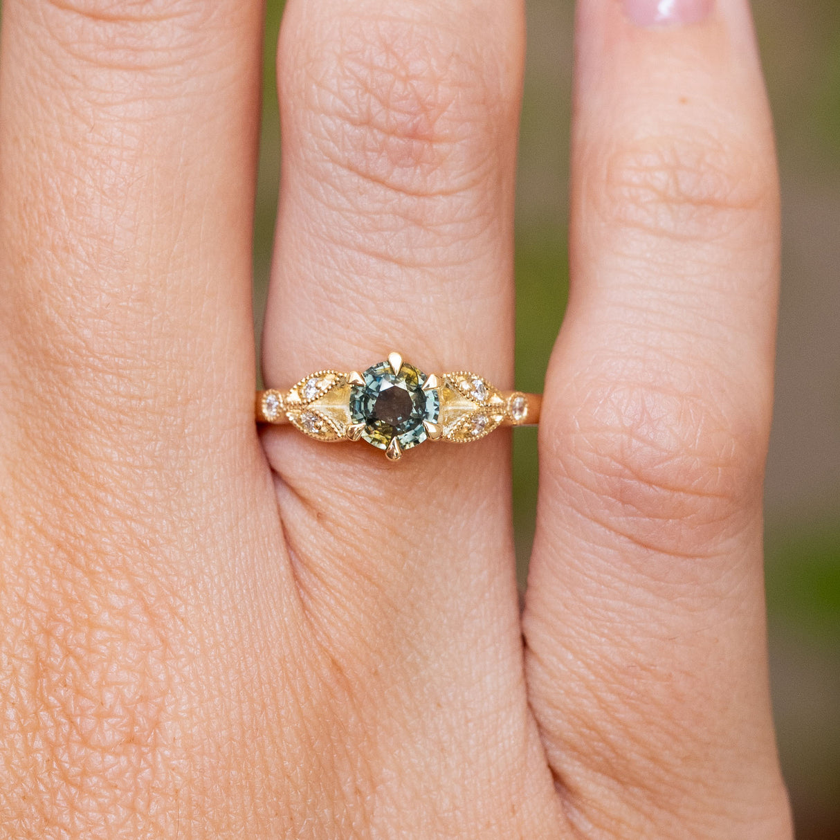 A close-up of a hand wearing a vintage-style yellow gold engagement ring with a central round green sapphire and small side diamonds.