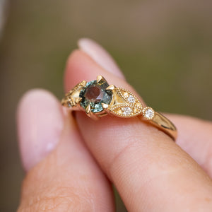 A close-up of a Vintage-inspired engagement ring with a central round green sapphire and small accent diamonds, with a yellow gold band worn on a finger
