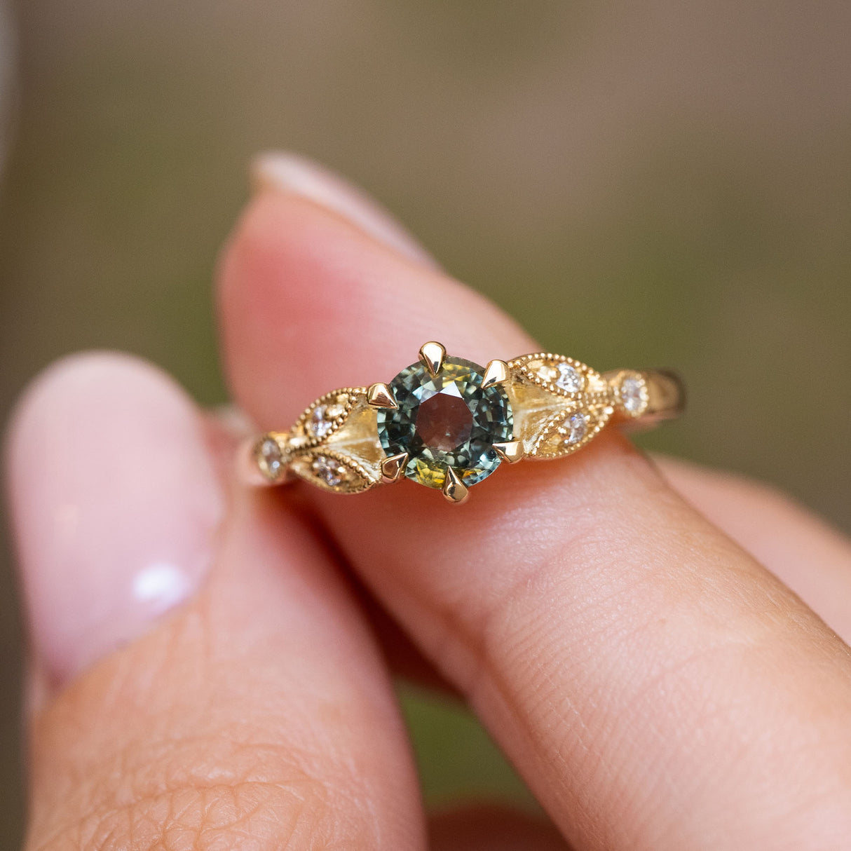 A close up of a Vintage-inspired, Green sapphire and diamond engagement ring photographed held between two fingers against a blurred indoor setting.