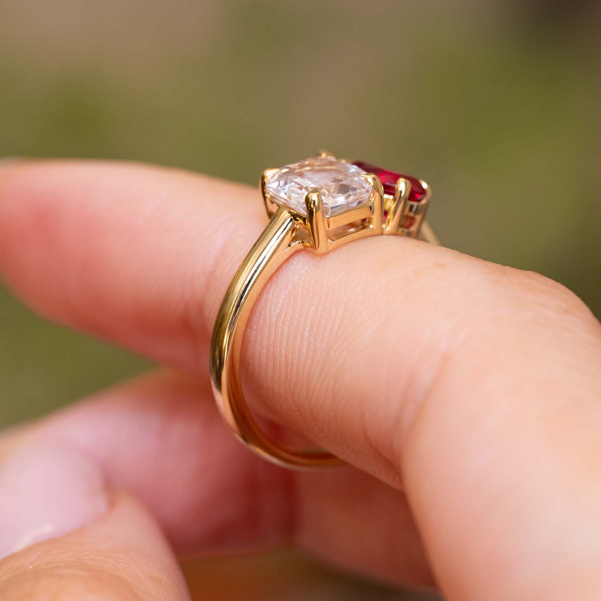 A close-up shot of a yellow gold Antique-inspired, Pink sapphire and spinel Toi et Moi engagement ring seen from the side worn on a finger against a blurred background.