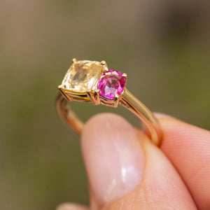 A close-up of an Antique-inspired, Yellow and pink sapphire Toi et Moi engagement ring in yellow gold and claw settings, hald between the fingers against a blurred green background.