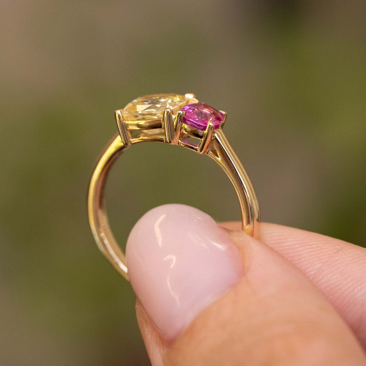 A close-up of a gold ring held between fingers. The ring is an Antique-inspired, Yellow and pink sapphire Toi et Moi engagement ring in yellow gold, held up to show the profile of the ring