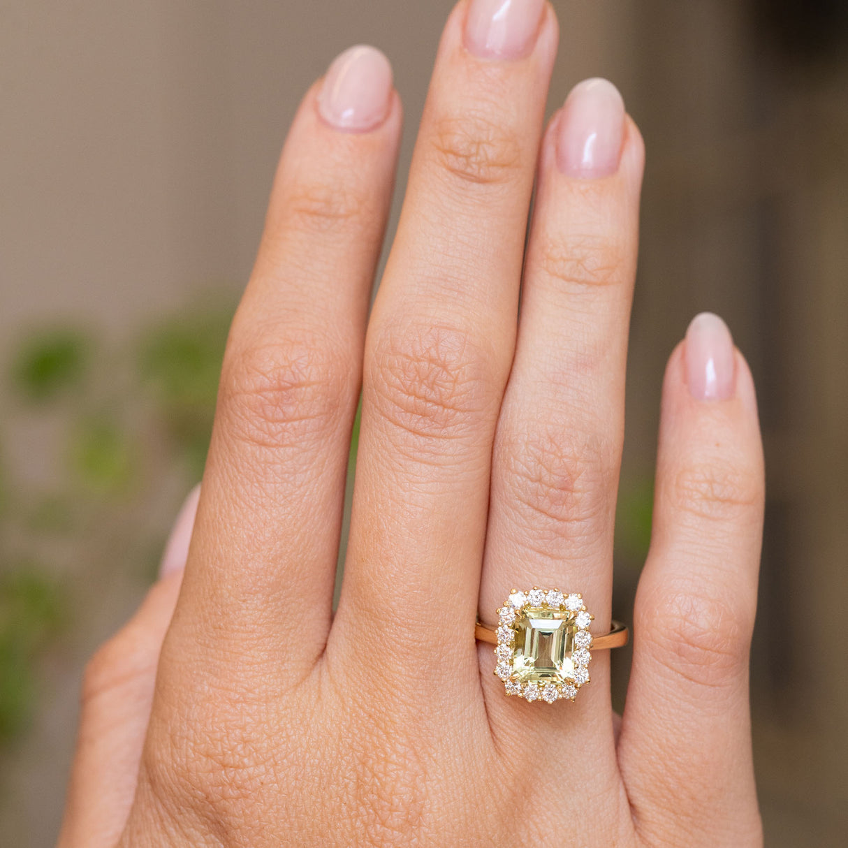 A close up of an Antique-inspired, Green beryl and diamond cluster engagement ring in yellow gold worn on a persons wedding finger. 