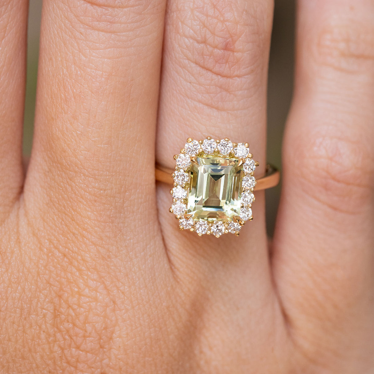 A close-up of a yellow gold engagement ring set with a large emerald-cut pale green beryl centre stone and a halo of small clear stones, worn on a finger. Antique-inspired.  