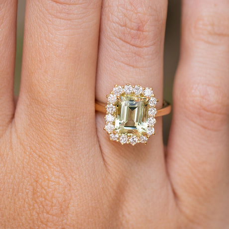 A close-up of a yellow gold engagement ring set with a large emerald-cut pale green beryl centre stone and a halo of small clear stones, worn on a finger. Antique-inspired.  