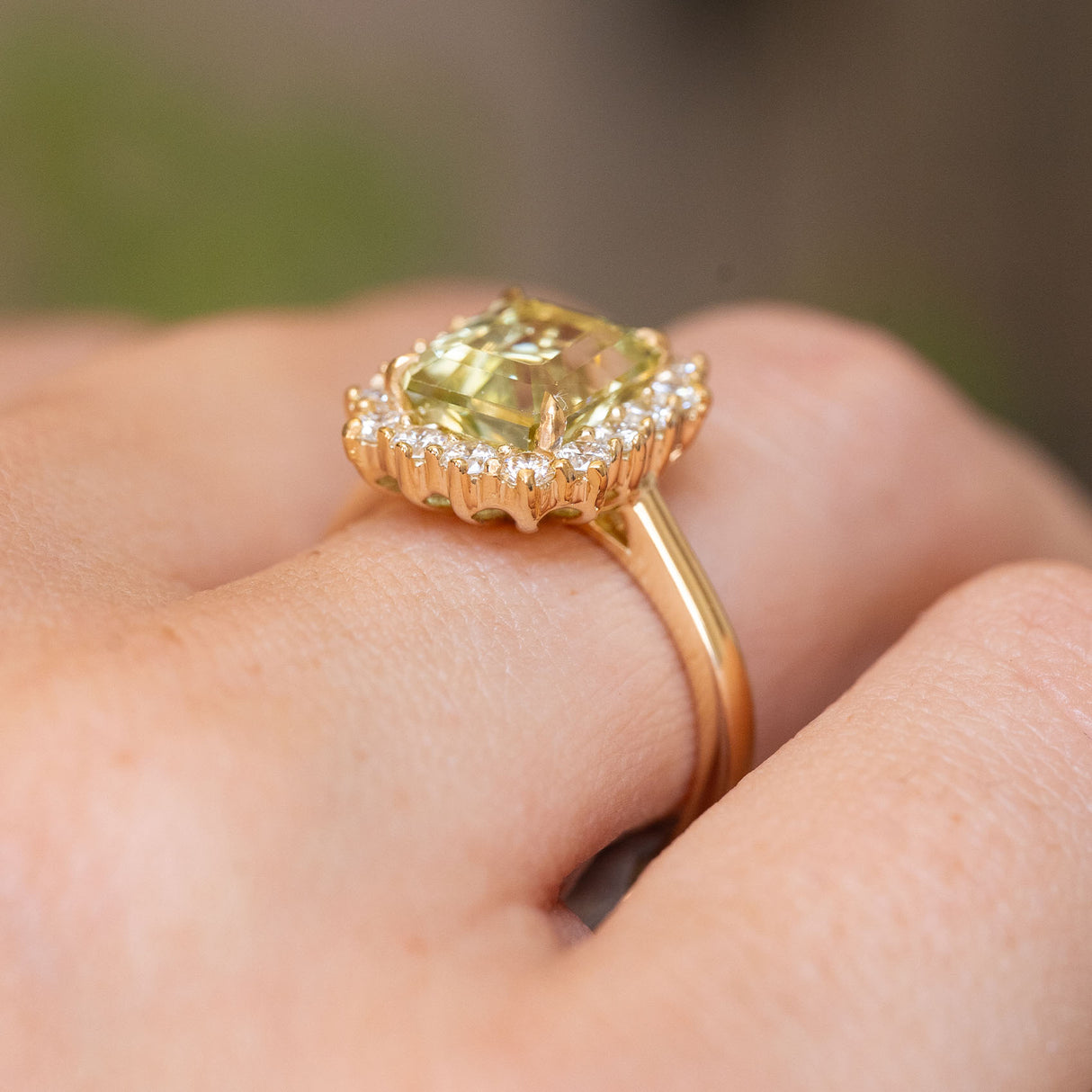 A close up of the low profile and basket setting of an Antique-inspired, Green beryl and diamond cluster engagement ring with a yellow gold band, worn on a persons finger. 