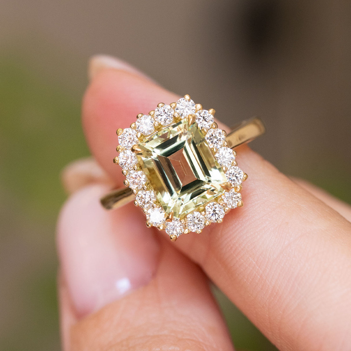 A close-up of the central emerald-cut green beryl and surrounding cluster of diamonds on an Antique-inspired, yellow gold cluster engagement ring, worn on a person's finger against a soft blurred background.