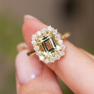 A close-up of the central emerald-cut green beryl and surrounding cluster of diamonds on an Antique-inspired, yellow gold cluster engagement ring, worn on a person's finger against a soft blurred background.