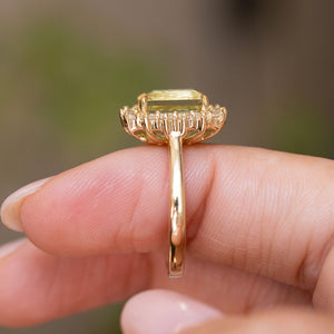 A close up of the basket setting and yellow gold band of an Antique-inspired, Green beryl and diamond cluster engagement ring worn on a finger against a softly blurred background. 