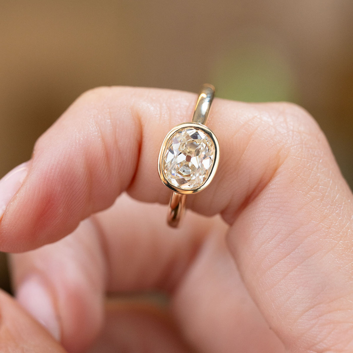 Antique-inspired elongated cushion diamond engagement ring held between the fingers, showing the warm gold bezel and distinctive old-mine facets.