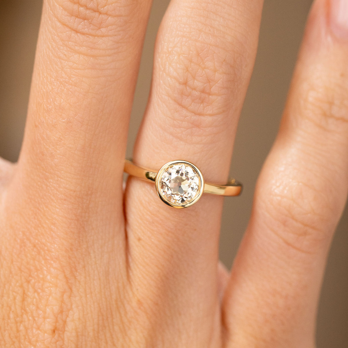 An up close picture of a Antique-inspired, Old-cut diamond solitaire engagement ring with a yellow gold basil rub over setting worn on a finger.
