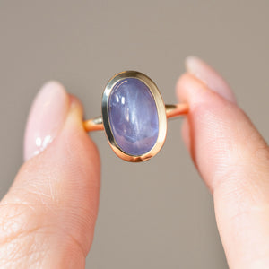 Detailed photograph of the oval cabochon sapphire held between fingers, highlighting its lavender-blue hues and natural cat's eye sheen.