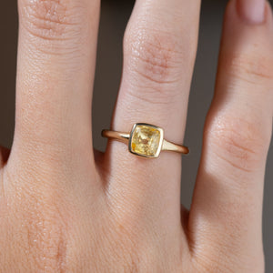 Close-up of the square cushion-cut yellow sapphire ring on a finger, showing its golden reflections and smooth bezel setting.