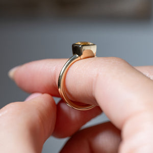 Side profile of the yellow gold solitaire ring held between fingers, revealing the elevated square bezel and polished band.
