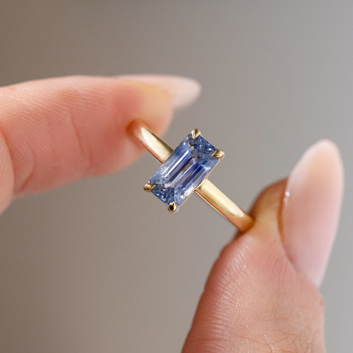 Detailed close-up of the elongated blue sapphire solitaire held between fingers, highlighting its crisp geometric facets and elegant four-claw setting.