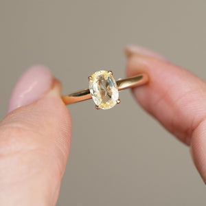 Detailed close-up of the oval-cut sapphire ring held between fingertips, emphasising its delicate lemon-yellow colour and polished gold band.