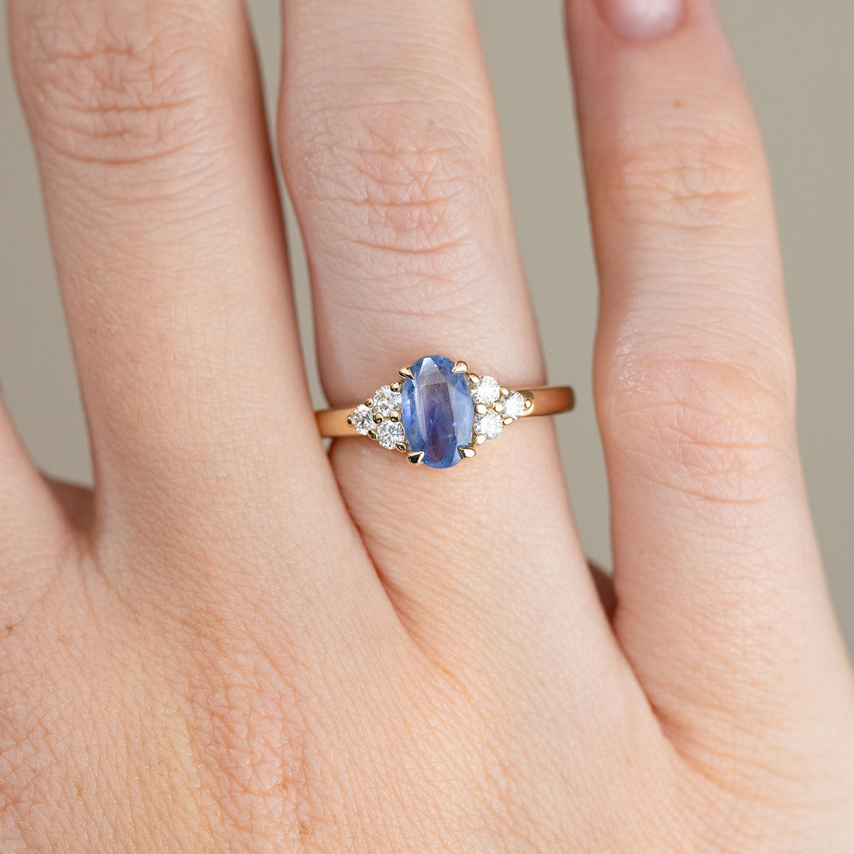 Close-up of an oval blue sapphire engagement ring on a finger, with brilliant white diamonds arranged in trefoil clusters on either side.