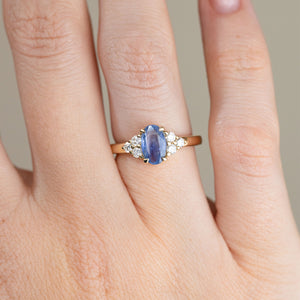 Close-up of an oval blue sapphire engagement ring on a finger, with brilliant white diamonds arranged in trefoil clusters on either side.