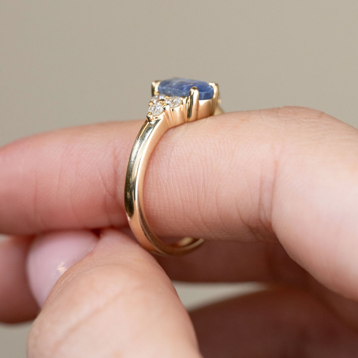 Profile photograph of the yellow gold engagement ring held between fingertips, displaying the sapphire's elevated claw setting and intricate diamond shoulders.