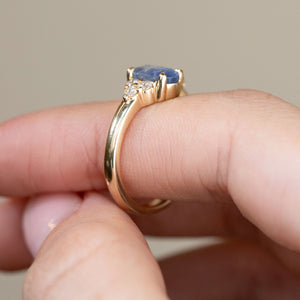 Profile photograph of the yellow gold engagement ring held between fingertips, displaying the sapphire's elevated claw setting and intricate diamond shoulders.