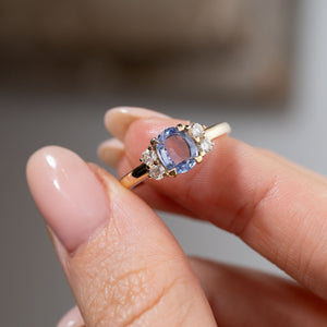 Close-up of the blue sapphire and diamond cluster engagement ring held between fingers, highlighting the sapphire's delicate blue reflections, surrounding diamonds and yellow gold claw setting.