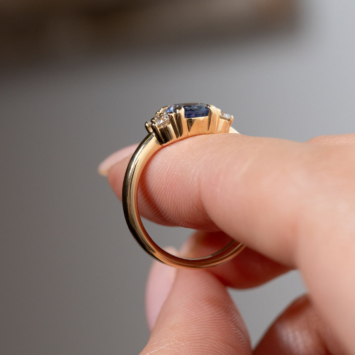 Side profile of the blue sapphire and diamond engagement ring held between fingers, revealing its raised gemstone settings, detailed claw arrangement and polished yellow gold band.