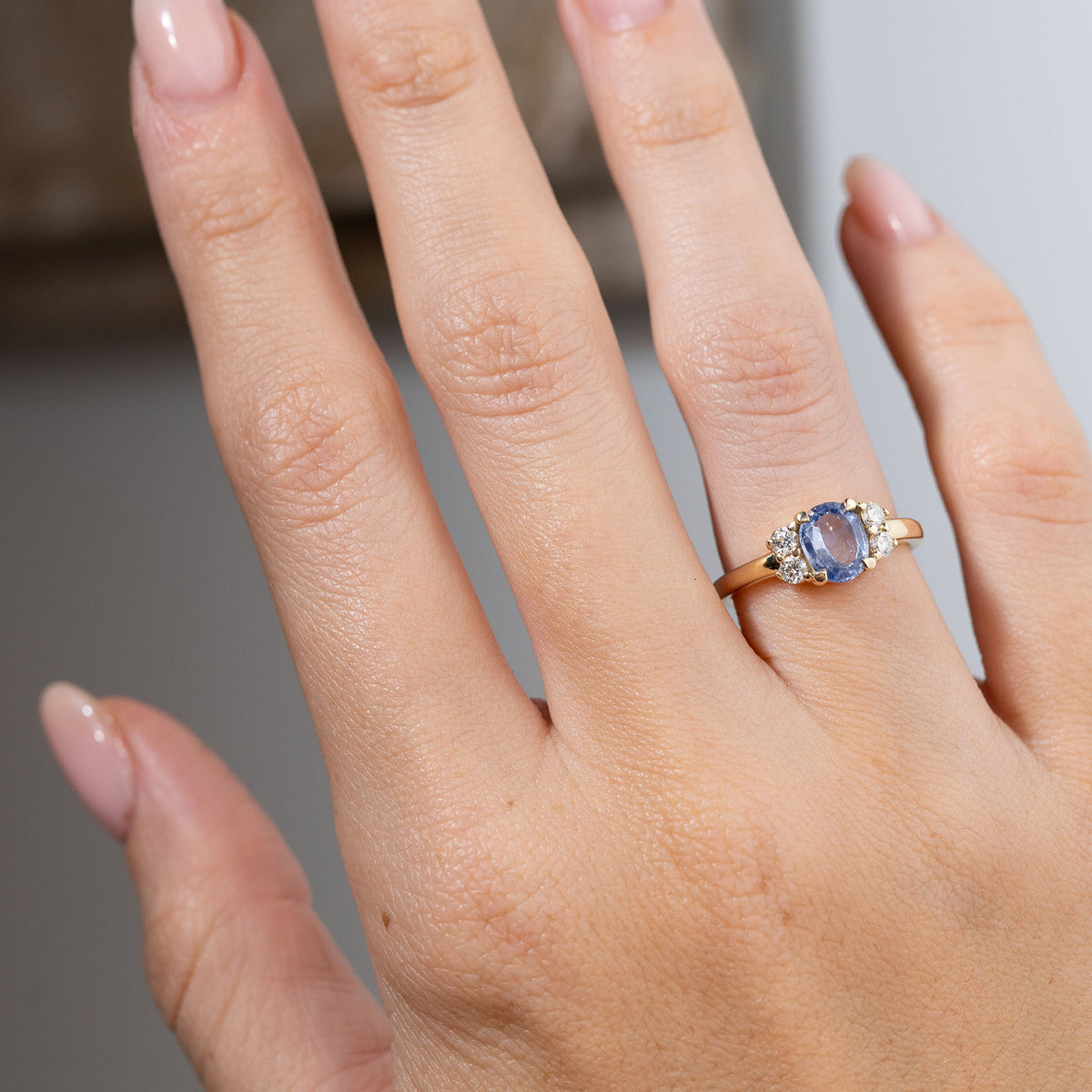 Vintage-inspired blue sapphire and diamond cluster engagement ring worn on a hand, displaying the oval-cut sapphire framed by two round diamonds on either side in yellow gold.