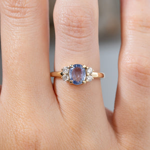 Close-up of the blue sapphire and diamond engagement ring worn on a finger, highlighting the pale blue oval gemstone, symmetrical diamond clusters and polished yellow gold setting.