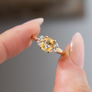 Detailed close-up of the vintage-inspired yellow sapphire and diamond engagement ring held between fingers, highlighting the cushion-cut gemstone, symmetrical diamond arrangement and polished gold claws.