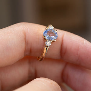 Hand-held close-up of the pastel blue sapphire engagement ring, showcasing the oval gemstone and sparkling diamond shoulders.