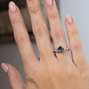 Sapphire and diamond engagement ring worn on a hand, featuring a dusky blue oval gemstone framed by decorative platinum shoulders.