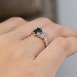 Angled side view of the engagement ring worn on a finger, revealing its raised sapphire setting, delicate diamond accents and detailed platinum shoulders.