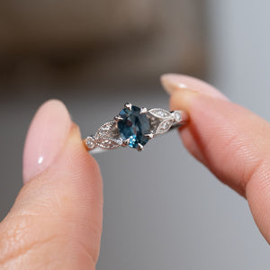 Detailed close-up of the vintage-inspired ring held between fingers, highlighting the sapphire's deep slate-blue tones and ornate laurel leaf-inspired diamond detailing.