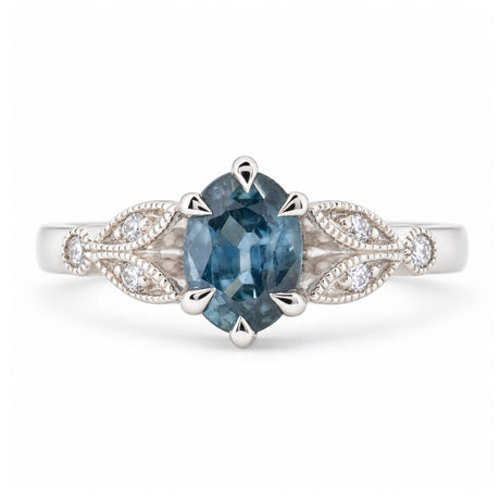 Front view of a vintage-inspired oval-cut sapphire engagement ring in platinum, featuring a muted blue gemstone and decorative diamond-set trefoil shoulders inspired by laurel leaves.