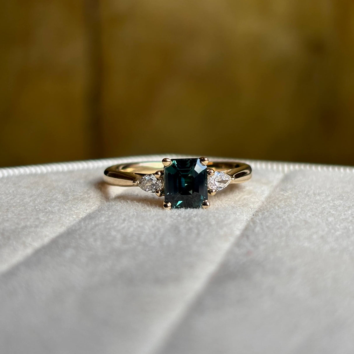 Green sapphire and diamond three-stone engagement ring on a blurred background showing colour of sapphire