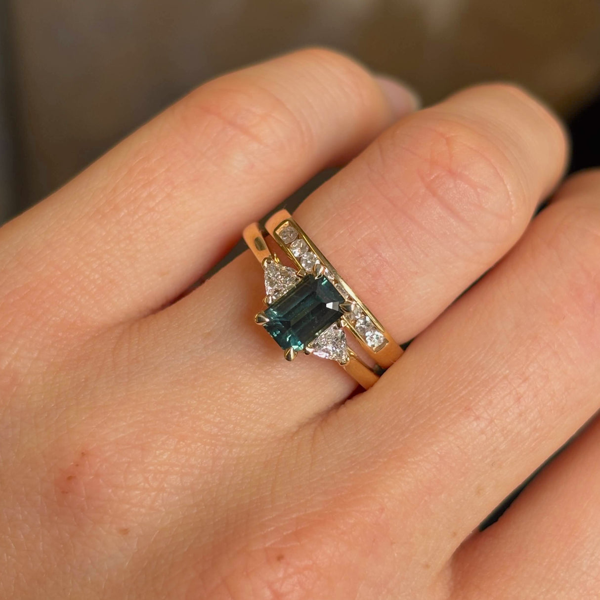 Green sapphire and diamond three-stone engagement ring on hand stacked with wedding band. 