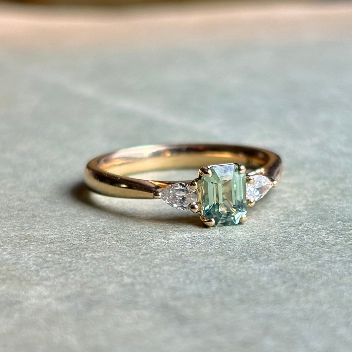 Green sapphire and diamond three-stone engagement ring resting on a neutral surface, showing the balance between the centre sapphire and diamond side stones.