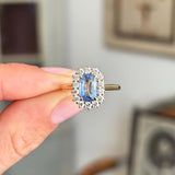 Hand holding a gold antique-inspired, blue elongated sapphire and diamond cluster engagement ring against a blurred indoor background