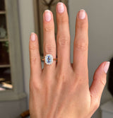An antique-inspired, blue sapphire and diamond cluster engagement ring in yellow gold worn on a hand against a blurred background.