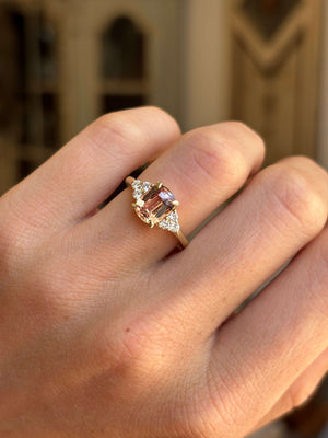 A close-up of a hand wearing a yellow gold ring with a central rectangular pink gemstone and small clear accent stones on the shoulders, set against a softly blurred indoor background.
