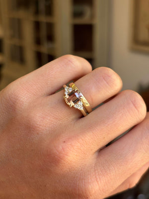 A vintage inspired yellow gold engagement ring with a central rectangular orange sapphire and smaller diamond accent stones, worn on a person's finger against a softly blurred indoor background.