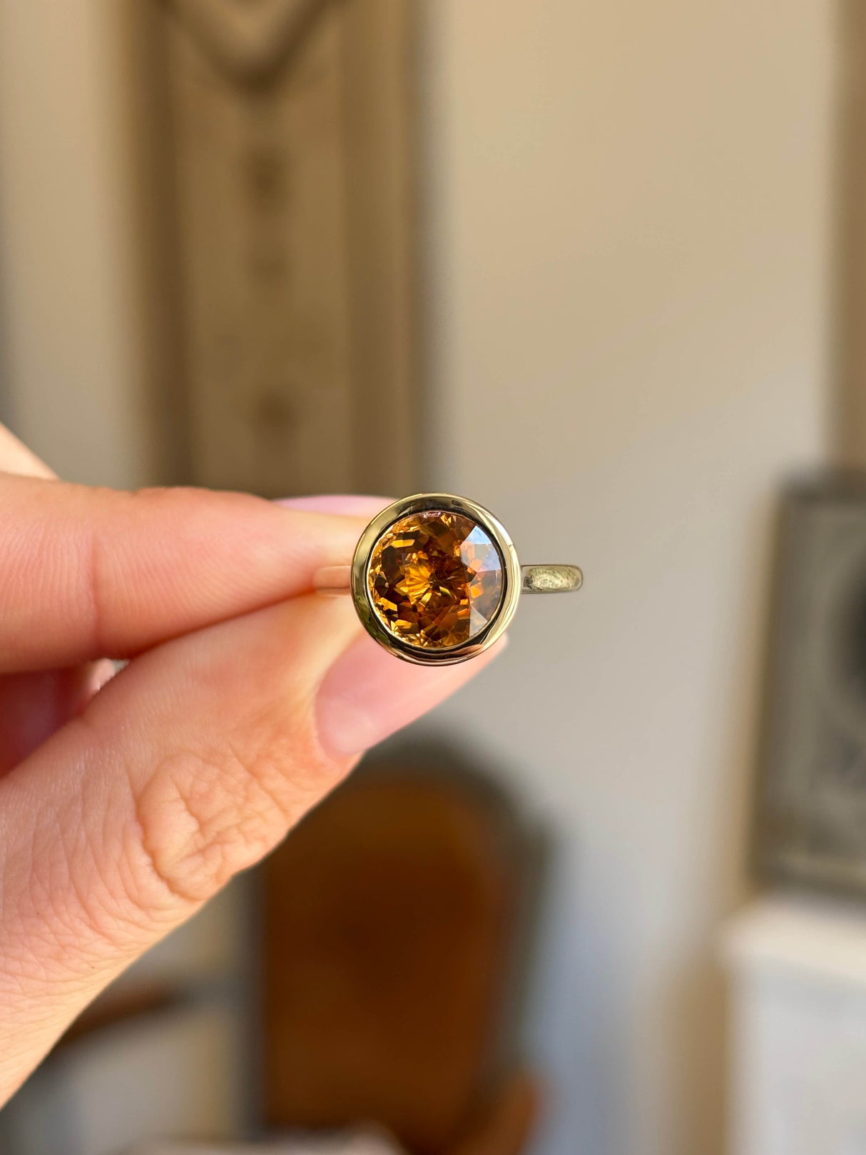 Antique-inspired, orange zircon solitaire ring with a bezel setting in yellow gold held between two fingers against an interior background