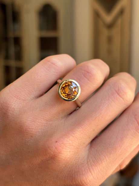 Closeup of an orange zircon ring in a bezel setting.  Antique-inspired and crafted in yellow gold.  Worn on a finger against a blurred interior setting. 