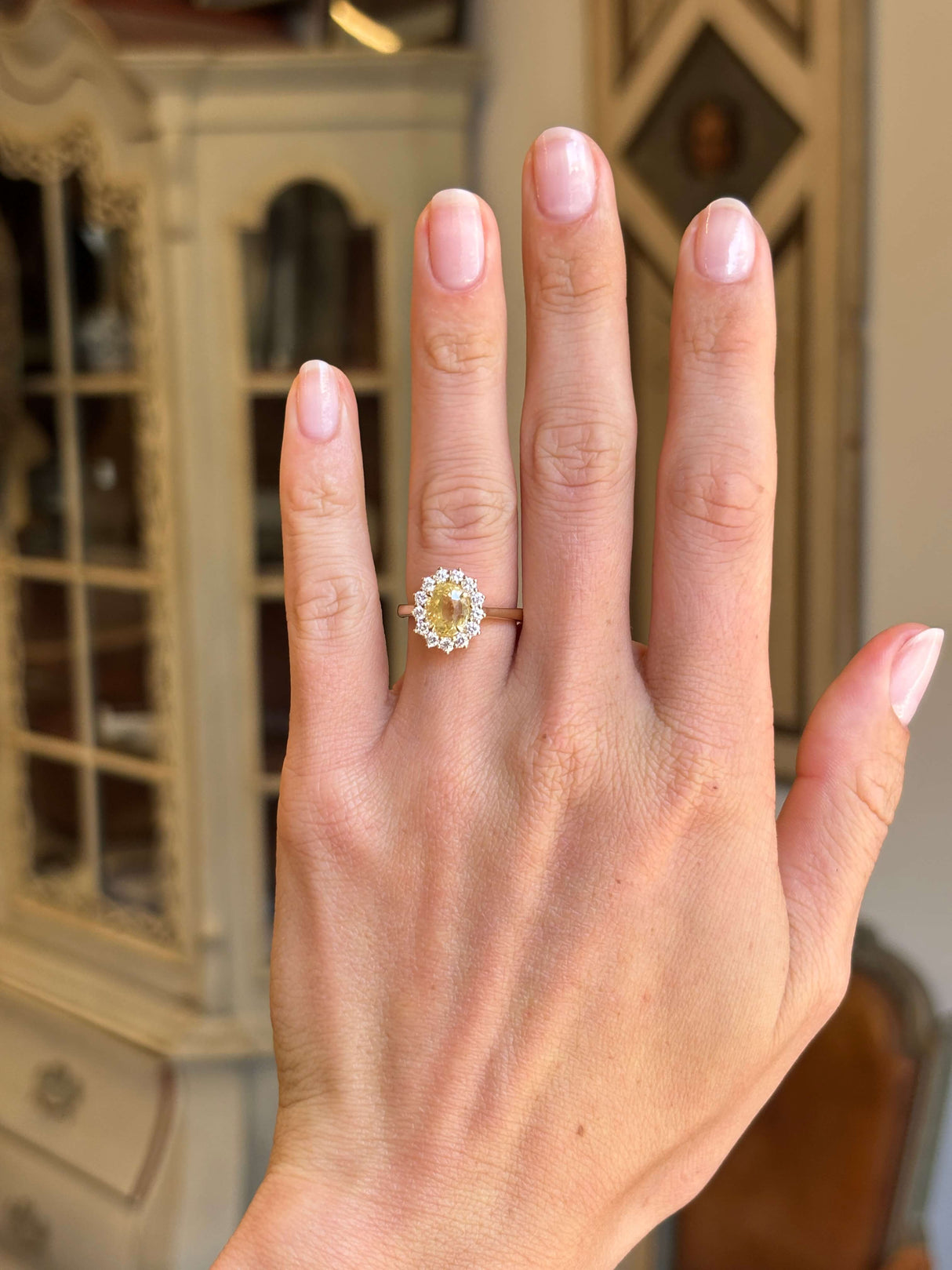 Antique-inspired, vibrant yellow sapphire and diamond cluster engagement ring in yellow gold worn on a hand against a blurred background. 