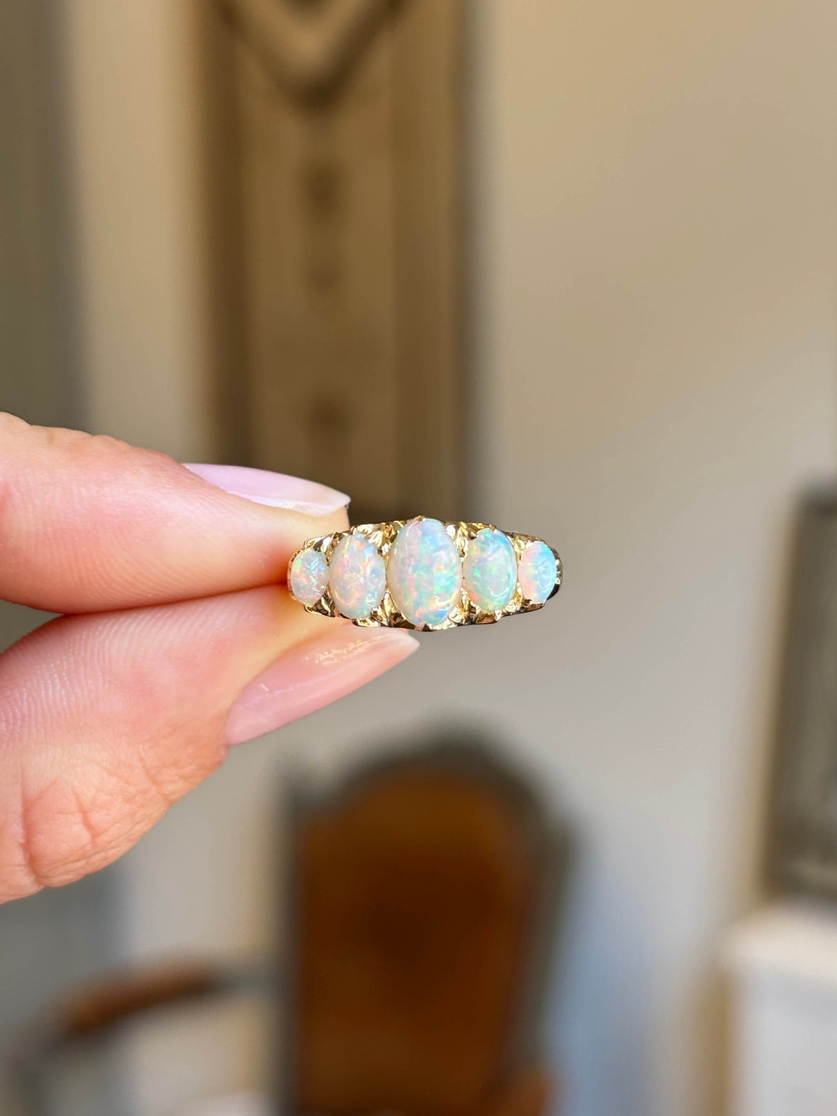 A close-up of a hand holding an Antique, Victorian precious white opal five-stone ring, with a softly blurred indoor background.