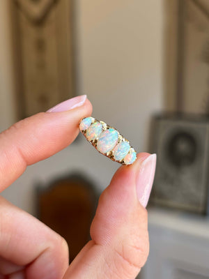 A close-up of a hand with holding an Antique, Victorian gold ring set with five iridescent opal stones against a blurred indoor background.