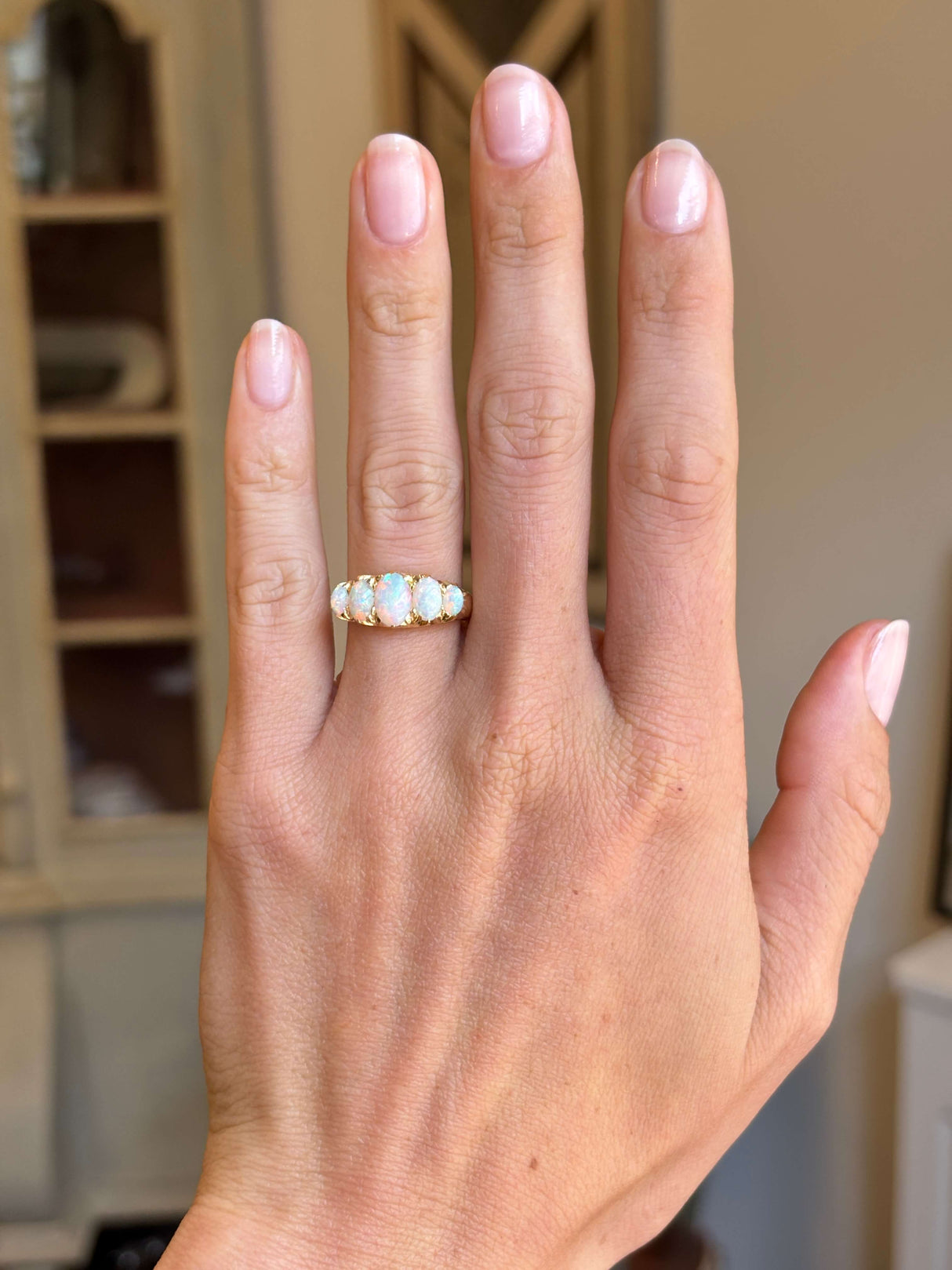 A close-up of a hand wearing an Antique, Victorian precious white opal five-stone ring set in a gold band, with a softly blurred indoor background.