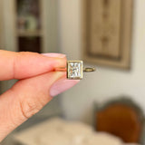 Vintage-inspired, diamond rub-over solitaire engagement gold ring held by a hand against a blurred indoor background