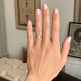 Hand wearing a Vintage-inspired, princess-cut diamond bezel solitaire engagement ring in gold on a blurred indoor background